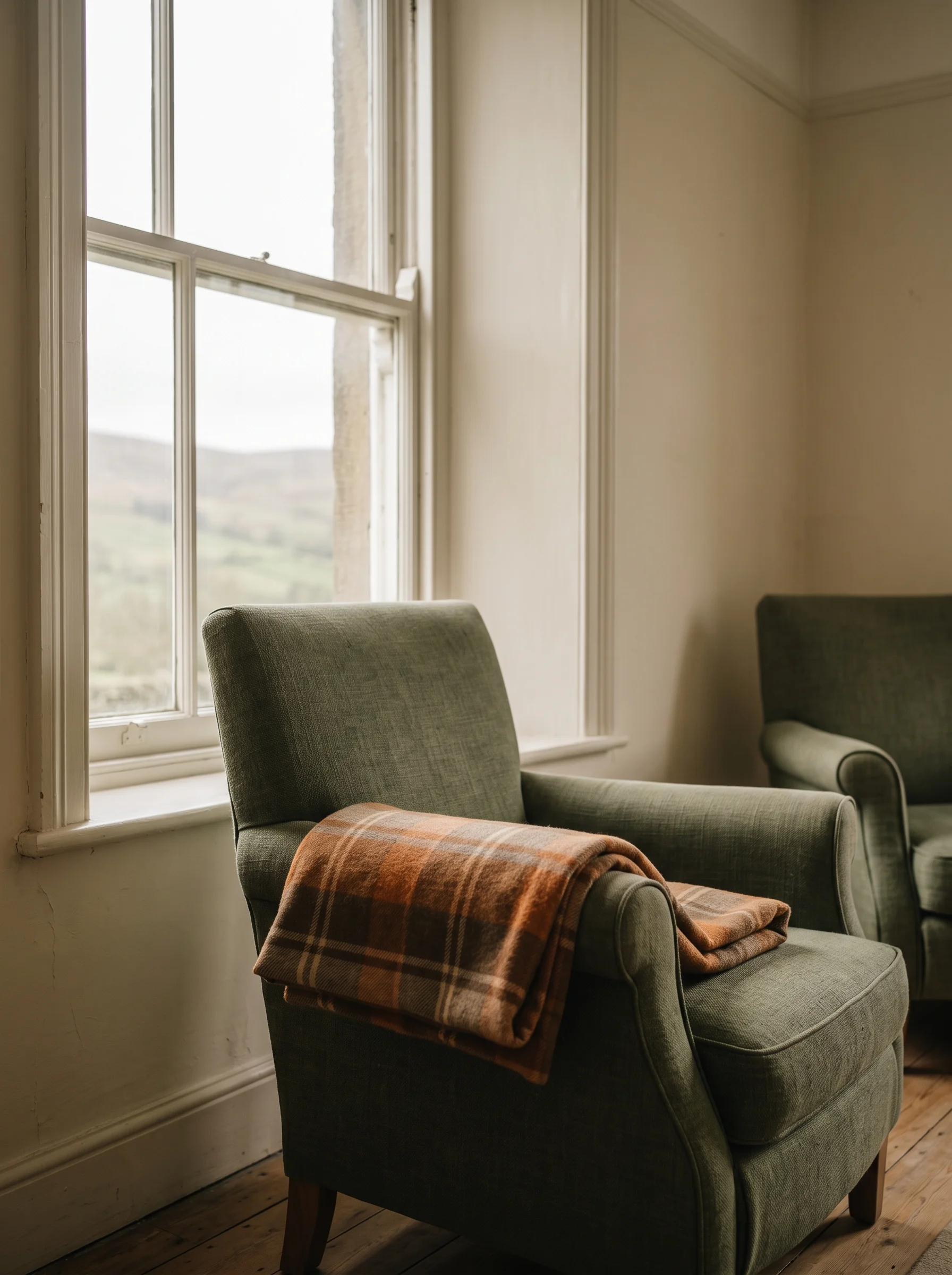 An armchair beside a sash window with a folded blanket on its arm, grey hill light across the seat